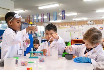 Group of young students in lab coats conduct a hands‑on science experiment at a classroom table using dropppers, containers and coloured liquids.