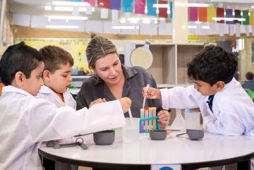 Students in lab coats work with a teacher at a classroom table, measuring and adding liquids into small containers as part of a guided science activity.
