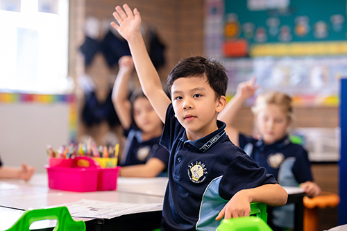 Student in a classroom sitting at a desk, with his hand in the air.