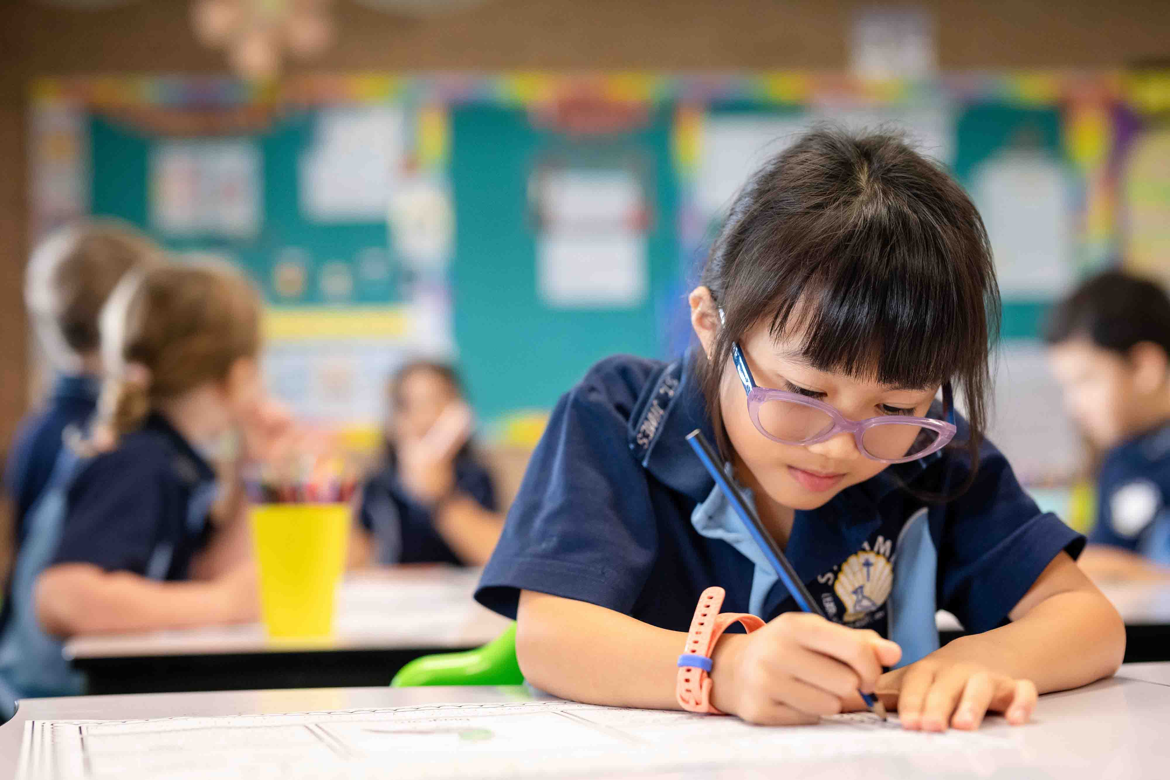 A girl wearing glasses is sitting a desk. She is holding a pencil and is writing.